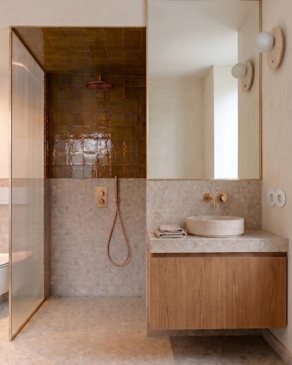 Texturas naturais e madeira encontram uma paleta neutra numa casa de banho pensada para o uso diário. Casa de banho com revestimento em pequenos azulejos, móvel de lavatório em madeira, bancada clara e duche ao fundo.