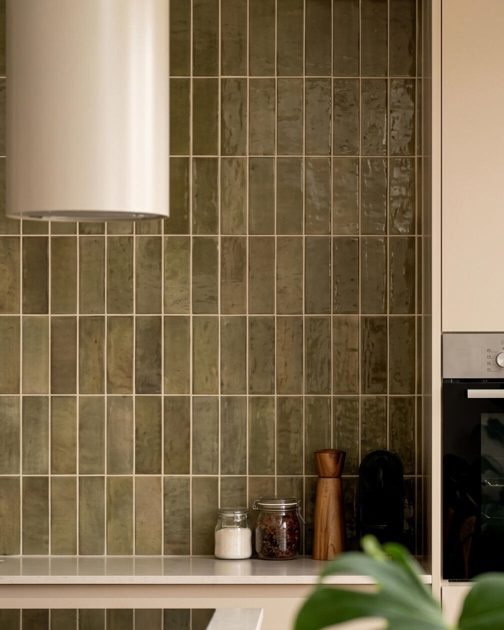 Um detalhe de cor e textura que acrescenta profundidade à composição da cozinha. Detalhe de cozinha com parede revestida por pequenos azulejos verdes de acabamento artesanal e armários em madeira clara.